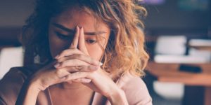 Woman with curly hair sitting with hands clasped in front of her face, expressing contemplation and emotional distress, reflecting themes of trauma and mental health support.